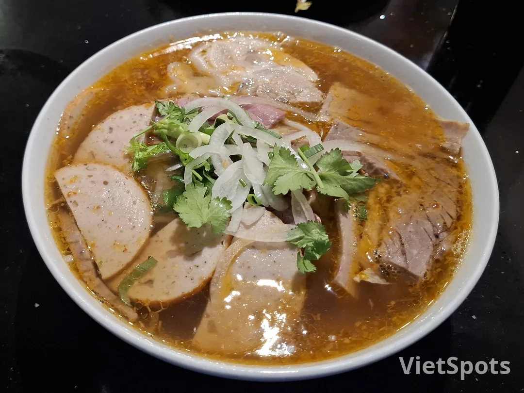 Tô bún bò Huế — nước dùng đỏ cam, thịt bò, chả cua, ngò hành trên
