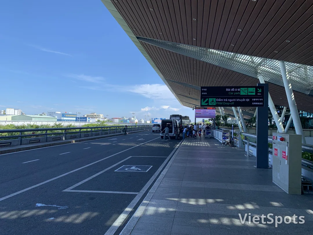 Cửa ra sân bay Đà Nẵng — mái gỗ hiện đại, biển hiệu Car park + PRM Drop-off tiếng Việt-Hàn-Anh-Trung, xe minivan đón khách
