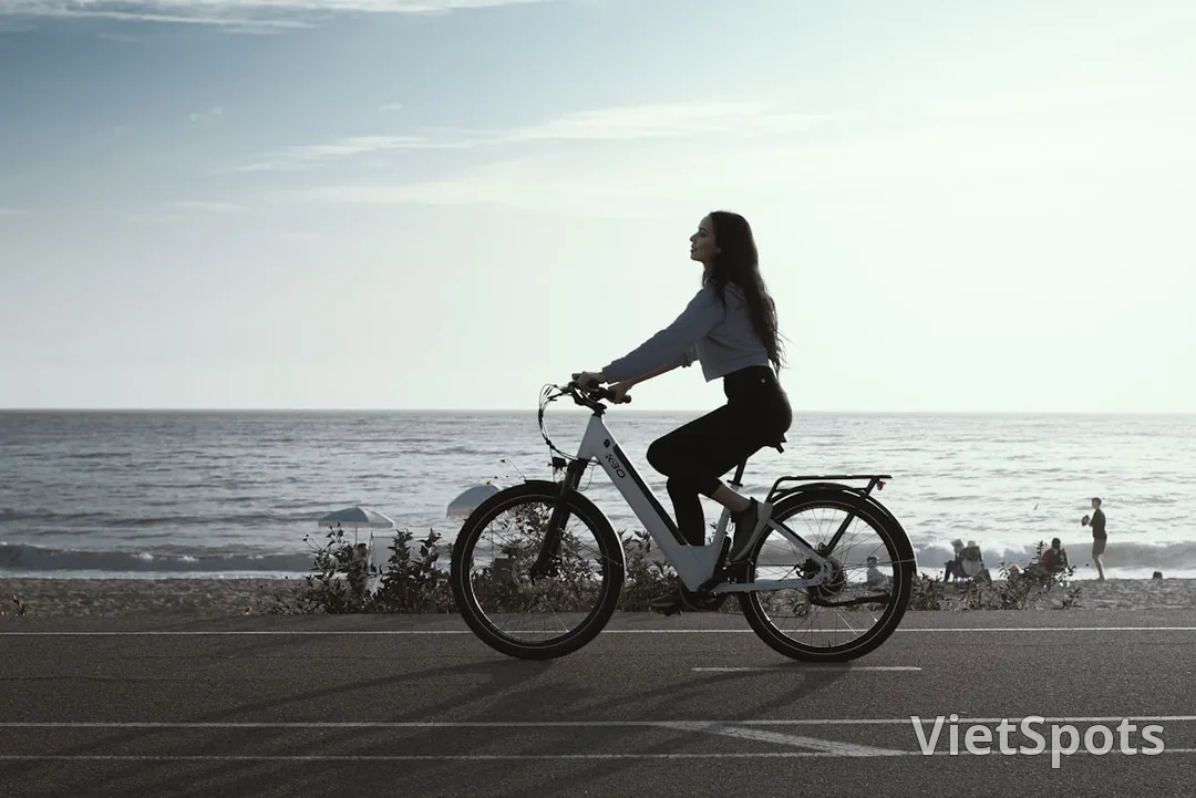 Em trẻ tóc dài mặc áo hoodie xám + quần đen casual, đạp xe city bike trắng trên đường ven biển, hoàng hôn nhẹ