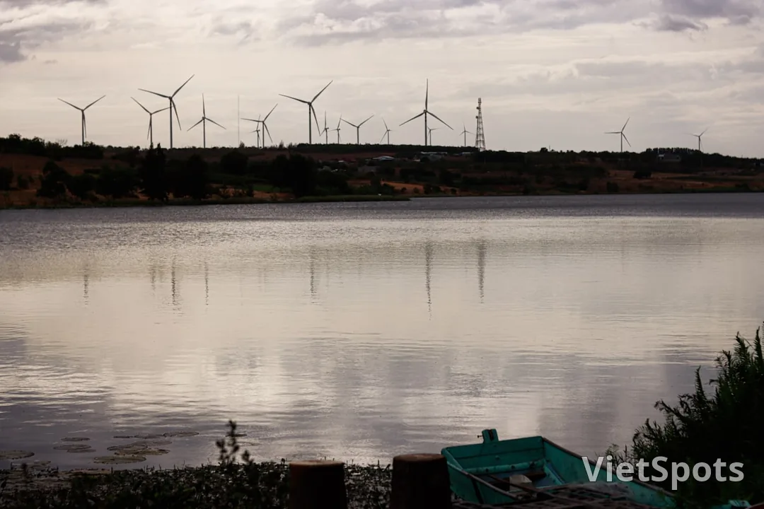 Hồ mặt phẳng lặng với hàng wind turbine phía xa, thuyền xanh nhỏ bên bờ