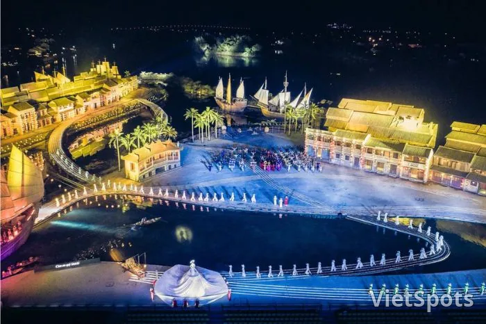 Aerial Ký Ức Hội An show 25.000m² — nhà cổ Hội An 2 bên, thuyền buồm, hàng cột trắng phun nước ánh sáng trên hồ, diễn viên áo dài đang biểu diễn giữa sân khấu