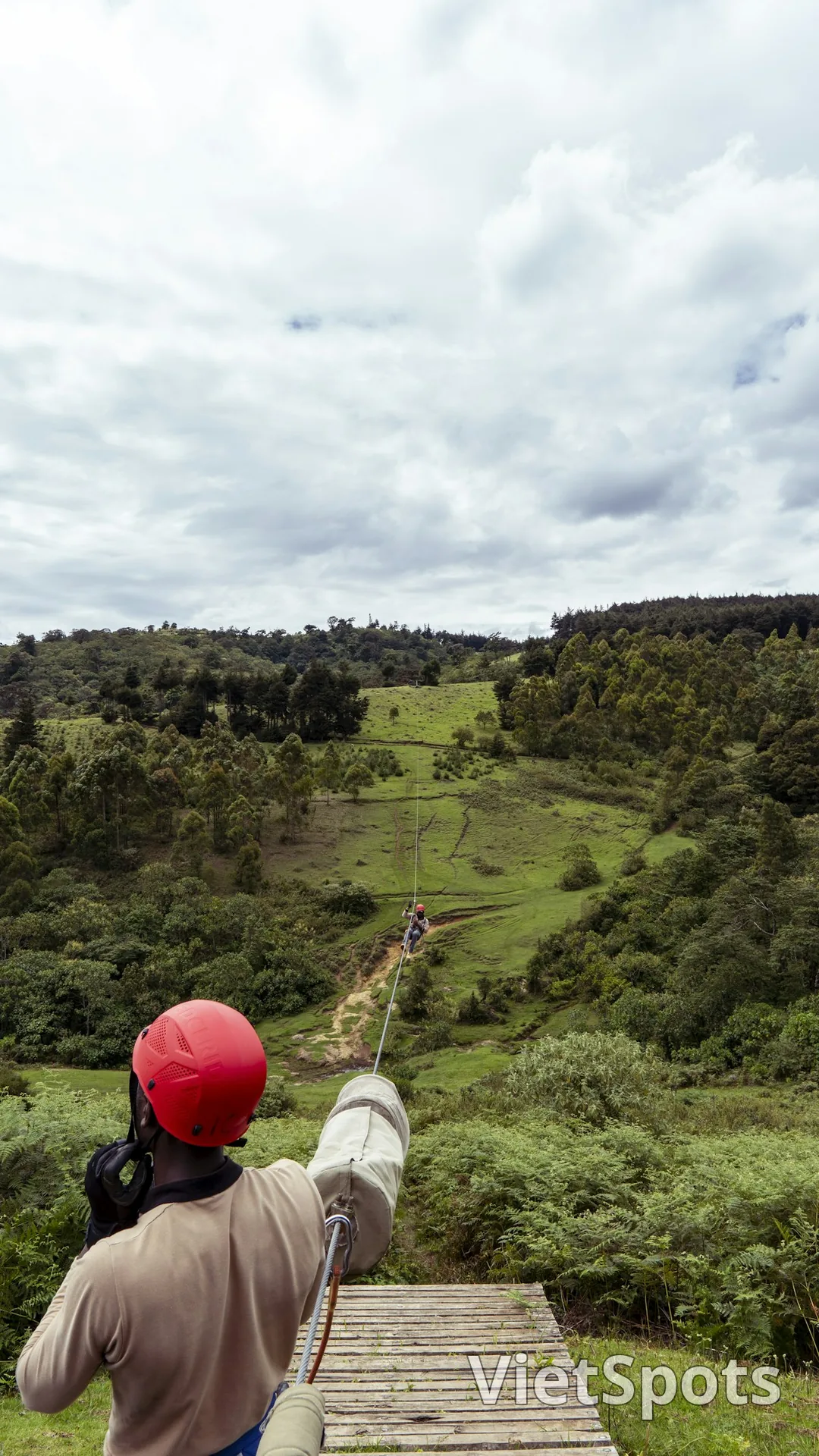 Người đội mũ bảo hiểm đỏ chuẩn bị trượt zipline xuống thung lũng xanh, dây zip line dài phía trước