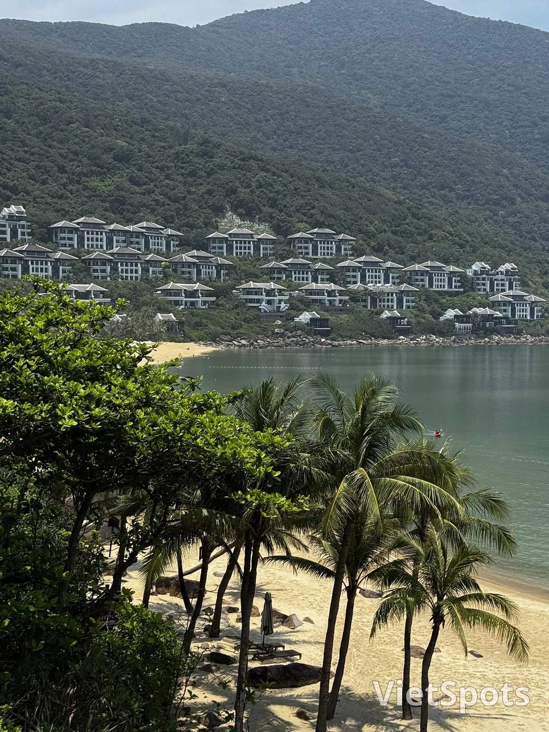 Các villa trắng xếp thành tầng trên sườn núi xanh, phía dưới là bãi biển và hàng dừa