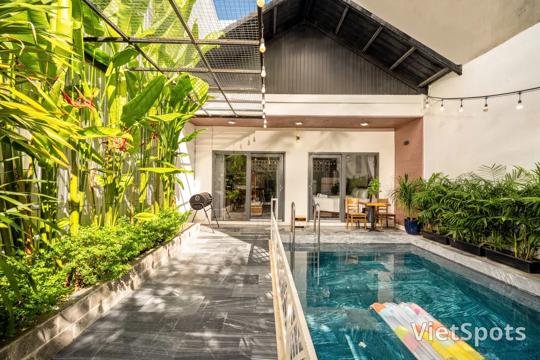 Sân trong của villa lúc nắng, hồ bơi hẹp chạy dọc sân, tường phủ chuối cảnh và thiên điểu, bàn hai ghế kê sát mép nước