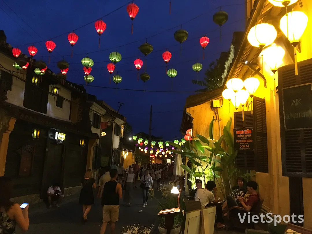 Ngõ phố cổ Hội An lúc chạng vạng, đèn lồng đỏ và xanh giăng ngang trên nền trời xanh thẫm, tường vàng hắt sáng ấm