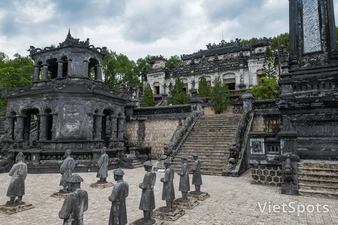 Sân chầu lăng Khải Định với hai hàng tượng quan bằng đá, phía sau là toà nhà bê tông xám đen chạm trổ dày đặc và bậc thang có rồng chầu