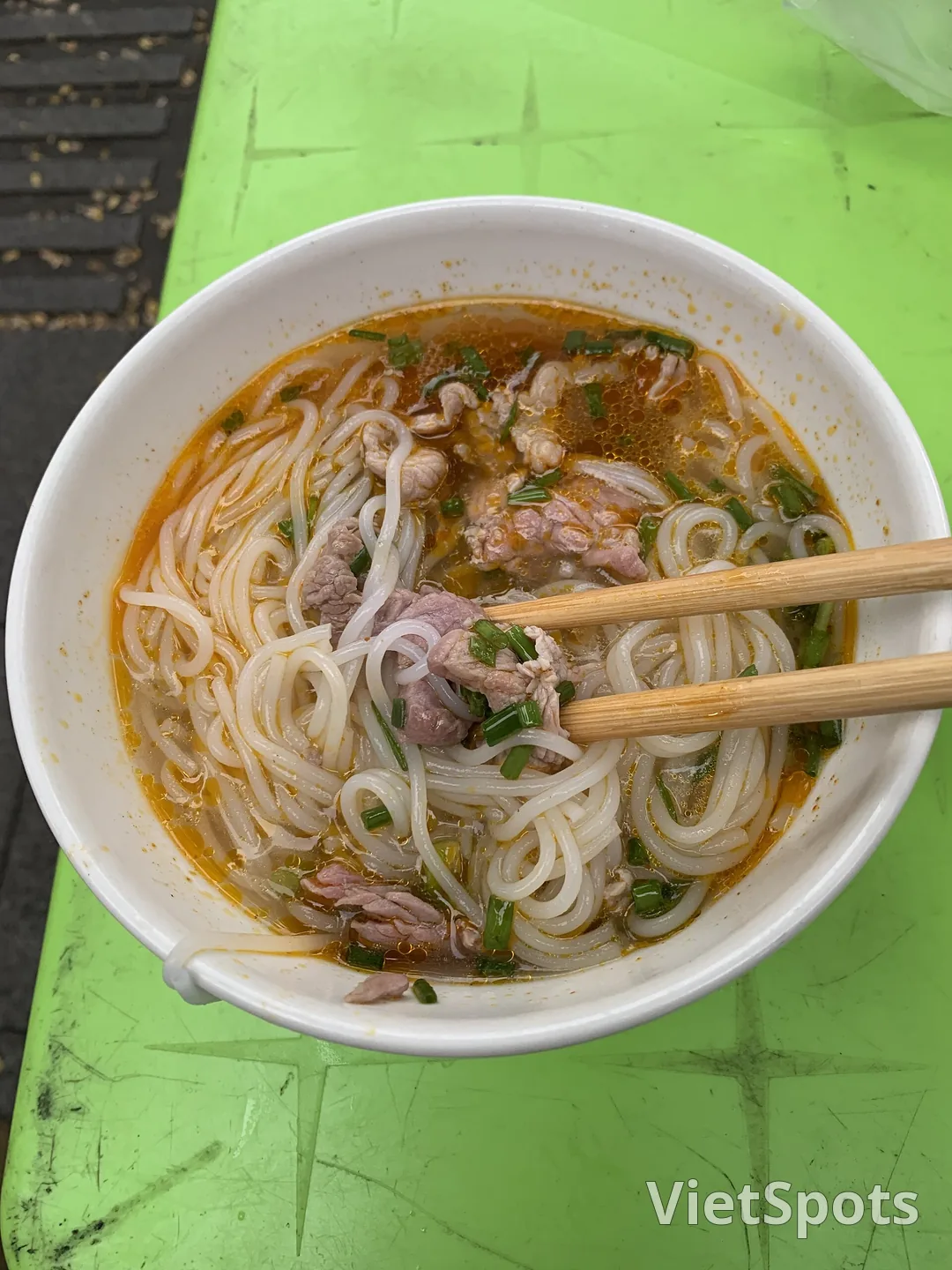 Tô bún bò Huế trên bàn nhựa xanh ở chợ Đông Ba, sợi bún tròn nhỏ, nước dùng nổi ớt màu, mấy lát thịt bò và hành lá