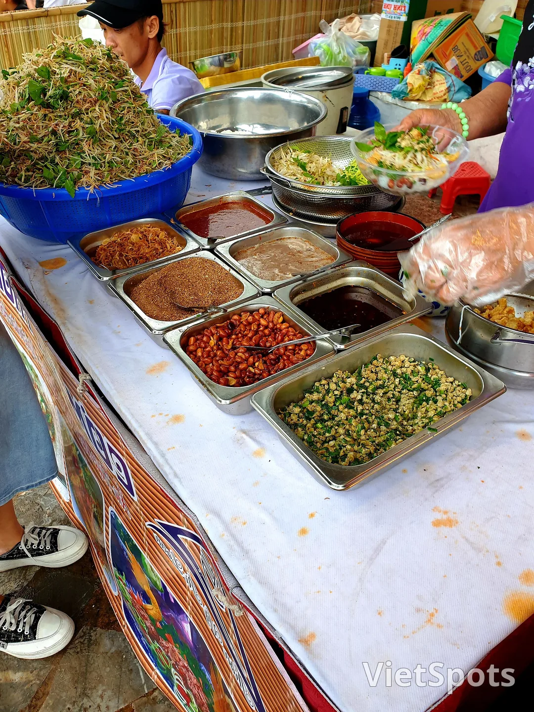 Sạp cơm hến bày cả chục khay thành phần: hến xào, ruốc, tóp mỡ, đậu phộng rang, mắm ớt, và một thúng rau trộn giá to phía sau