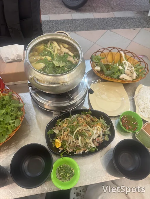 Nồi lẩu gà lá é bằng nhôm đặt trên bếp, lá é xanh nổi trong nước dùng, bên cạnh là rổ lá é tươi, đĩa gỏi gà và mấy chén muối ớt xanh
