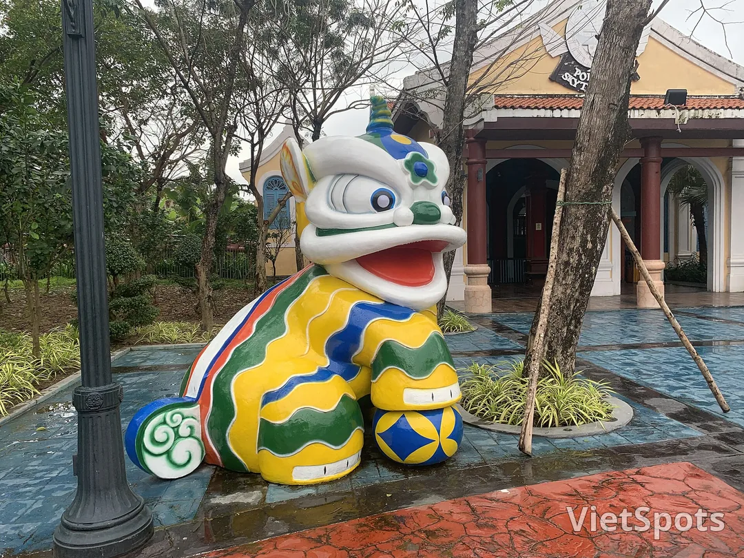 Tượng con lân sơn nhiều màu vàng xanh đỏ đặt trong khuôn viên công viên, phía sau là dãy nhà mái ngói