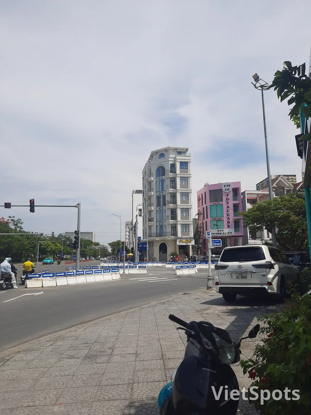 Vỉa hè Đà Nẵng với một chiếc xe máy dựng ở tiền cảnh, phía xa là ngã tư và vài người đi xe máy đội mũ bảo hiểm