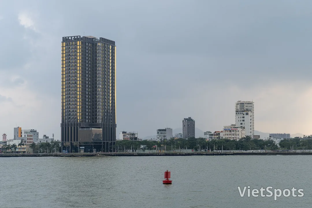 Bờ sông Hàn Đà Nẵng dưới trời xám, một toà tháp cao và dãy nhà thấp chạy dọc bờ, phao báo hiệu đỏ nổi trên mặt nước