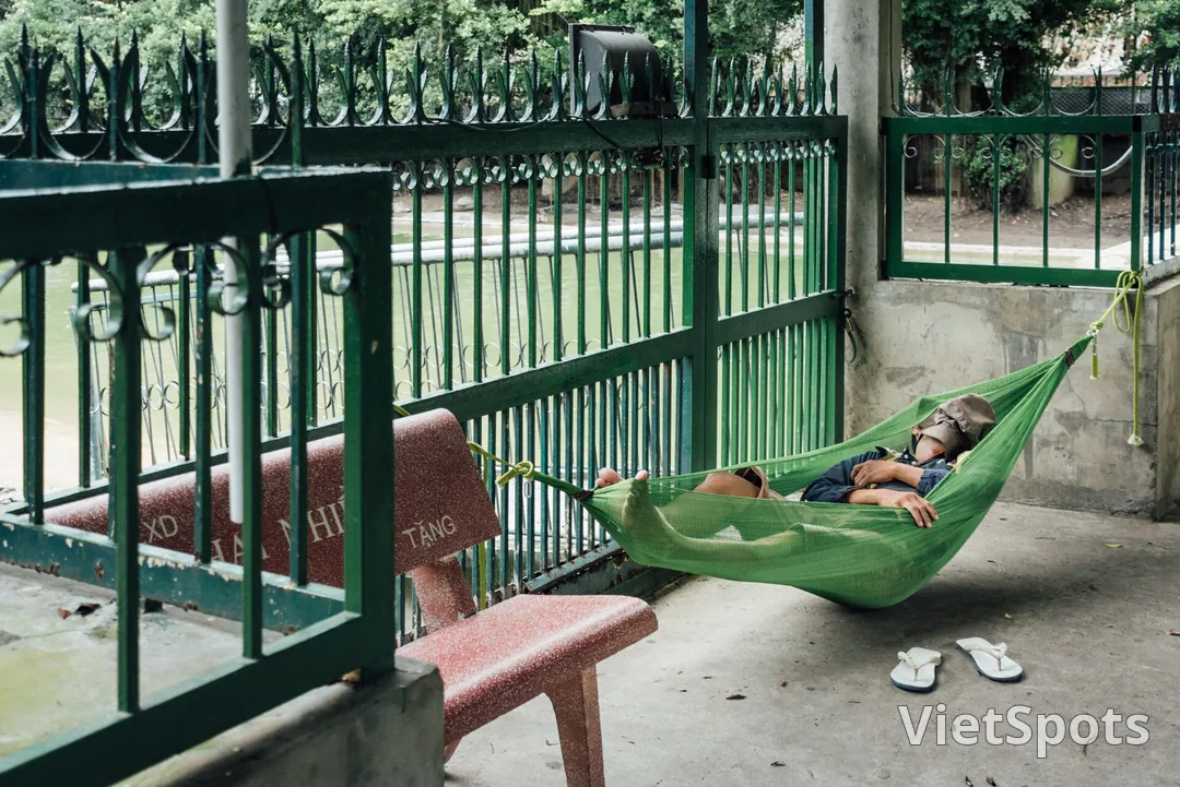 Một người đàn ông nằm ngủ trên chiếc võng lưới xanh mắc ngang lối đi bê tông, nón úp lên mặt, đôi dép bỏ lại dưới nền