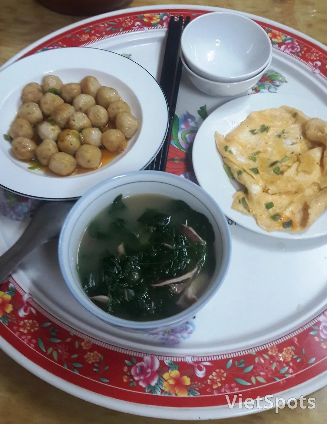 Mâm cơm khác trên khay viền đỏ: đĩa chả viên, trứng chiên, tô canh rau, đũa và chén nhỏ xếp chồng bên cạnh