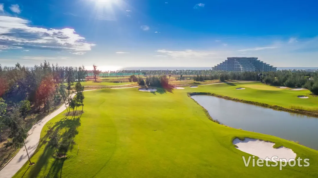 Một fairway rộng ở Vinpearl Golf Nam Hội An với hồ nước bên phải và rặng phi lao chắn gió ven biển