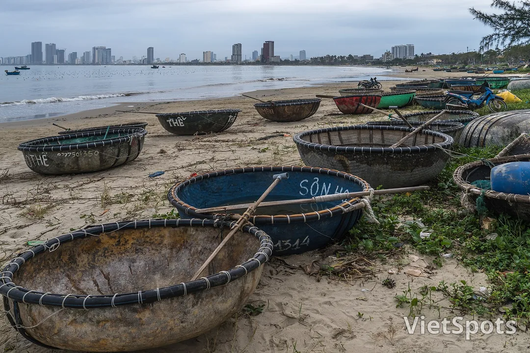 Hàng chục thuyền thúng kéo lên bãi cát ở Thọ Quang, trên vành thúng sơn tên chủ và số điện thoại, phía xa là dãy cao ốc bên kia vịnh