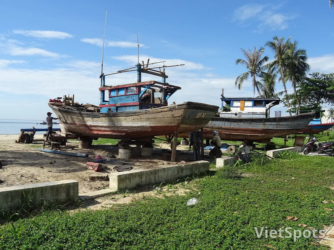 Hai chiếc tàu cá gỗ được kê lên đà ngay trên bãi để sửa, vài người đang làm việc dưới thân tàu, phía sau là hàng dừa