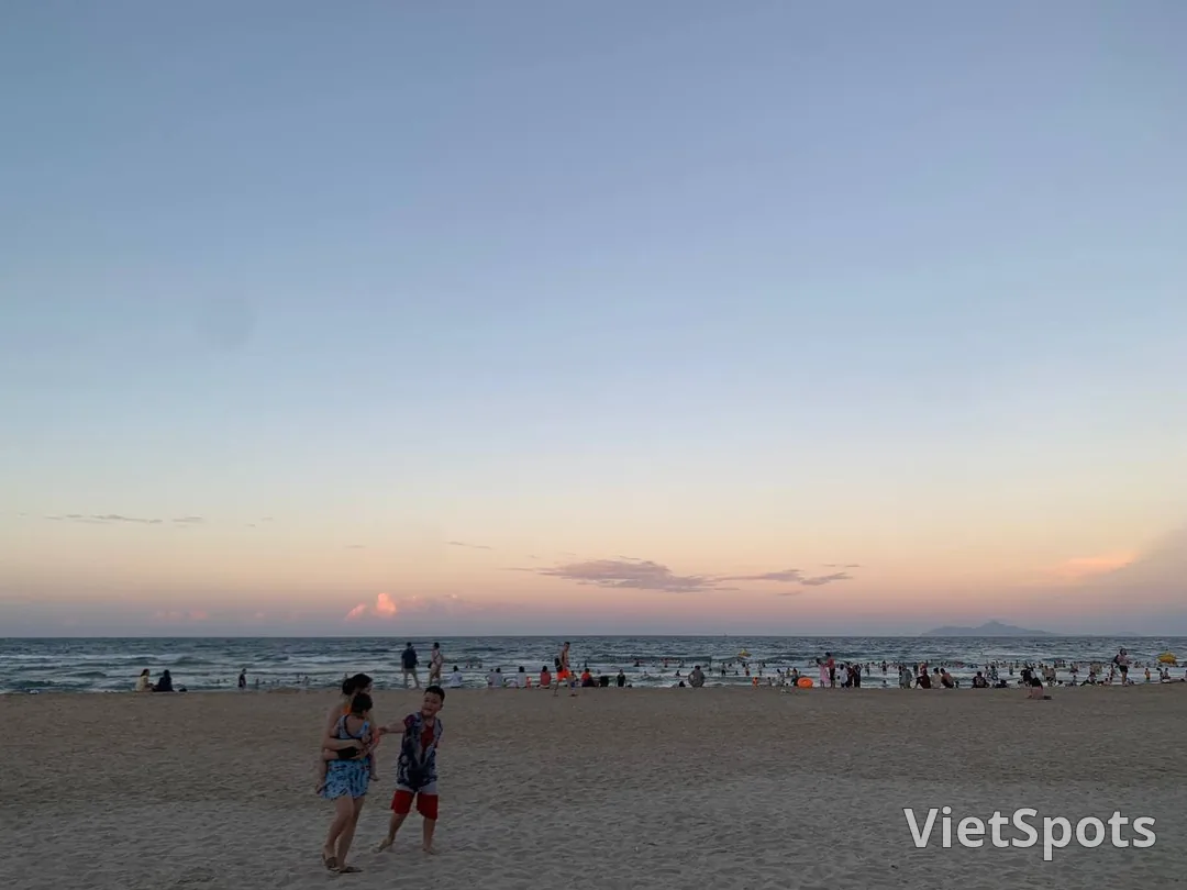 Bãi biển Đà Nẵng lúc chiều muộn, trời ngả hồng, hàng trăm người đang tắm dưới nước và đi lại trên cát