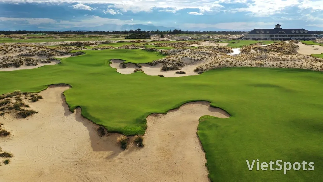 Một hố golf ven biển nhìn từ trên cao, fairway xanh len giữa những đụn cát hoang, nhà câu lạc bộ mái tối nằm ở phía xa