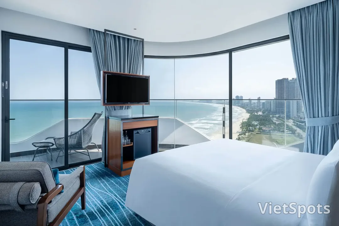Phòng khách sạn với cửa kính cong nhìn thẳng ra bãi biển Đà Nẵng, giường trắng và ghế mây ngoài ban công