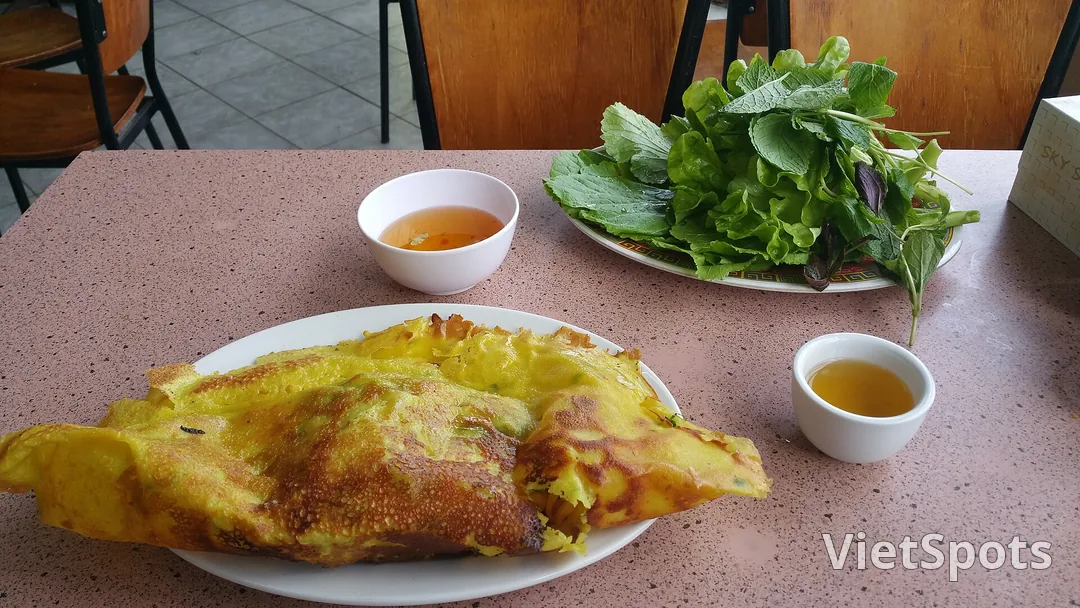 Chiếc bánh xèo vàng ruộm gập đôi trên đĩa, bên cạnh là đĩa rau sống xanh và hai chén nước chấm