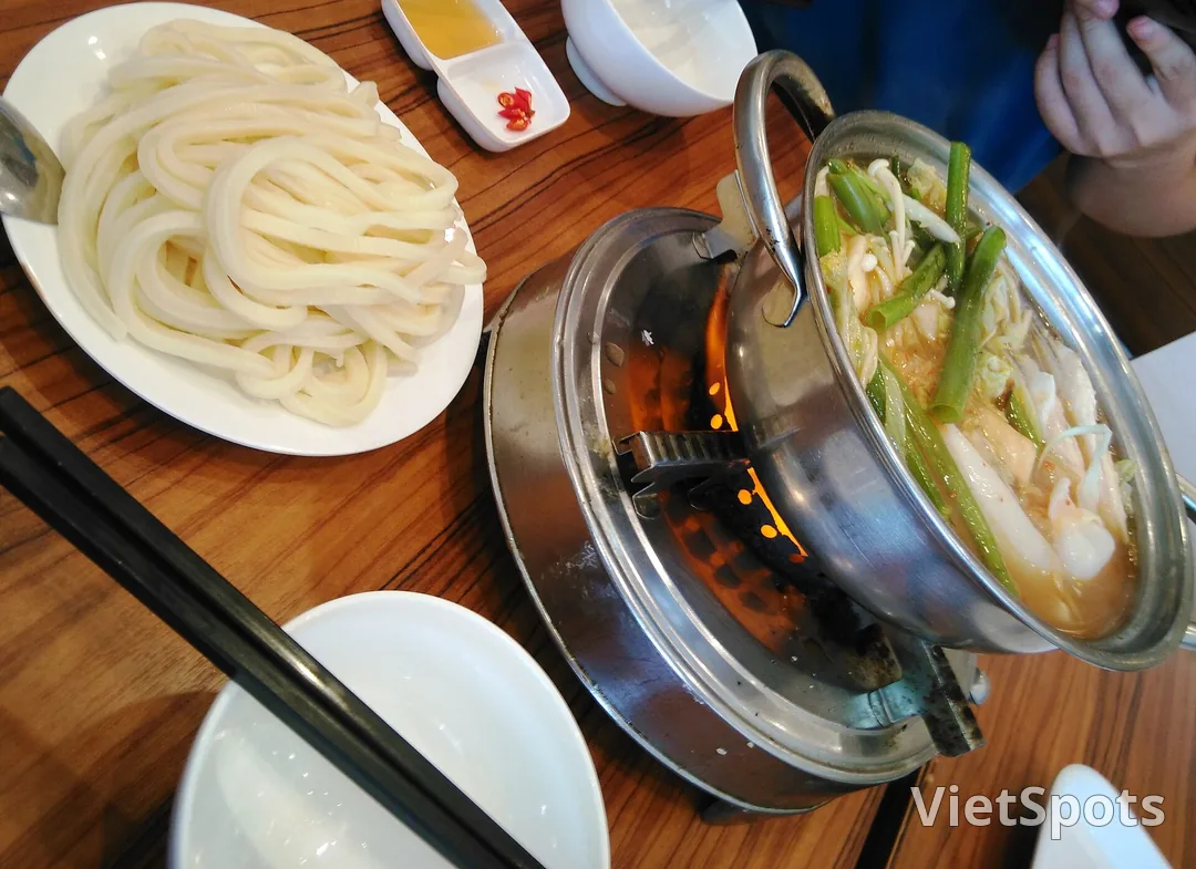 Nồi lẩu đang sôi trên bếp cồn với lửa cháy đỏ bên dưới, bên cạnh là đĩa bún trắng xếp cuộn