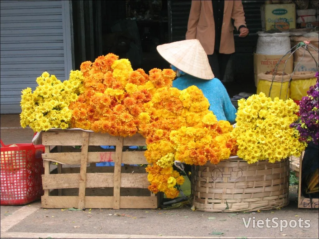 Sạp hoa vỉa hè với những bó cúc vàng và cam chất đầy trên thùng gỗ và giỏ tre, người bán đội nón ngồi phía sau