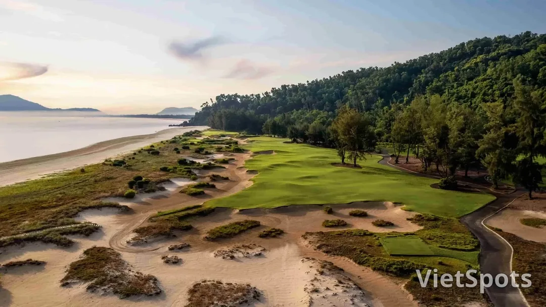 Fairway xanh giữa những đồi cát và bụi cỏ hoang ven biển, rừng phi lao phía sau, bên trái là bãi biển Lăng Cô