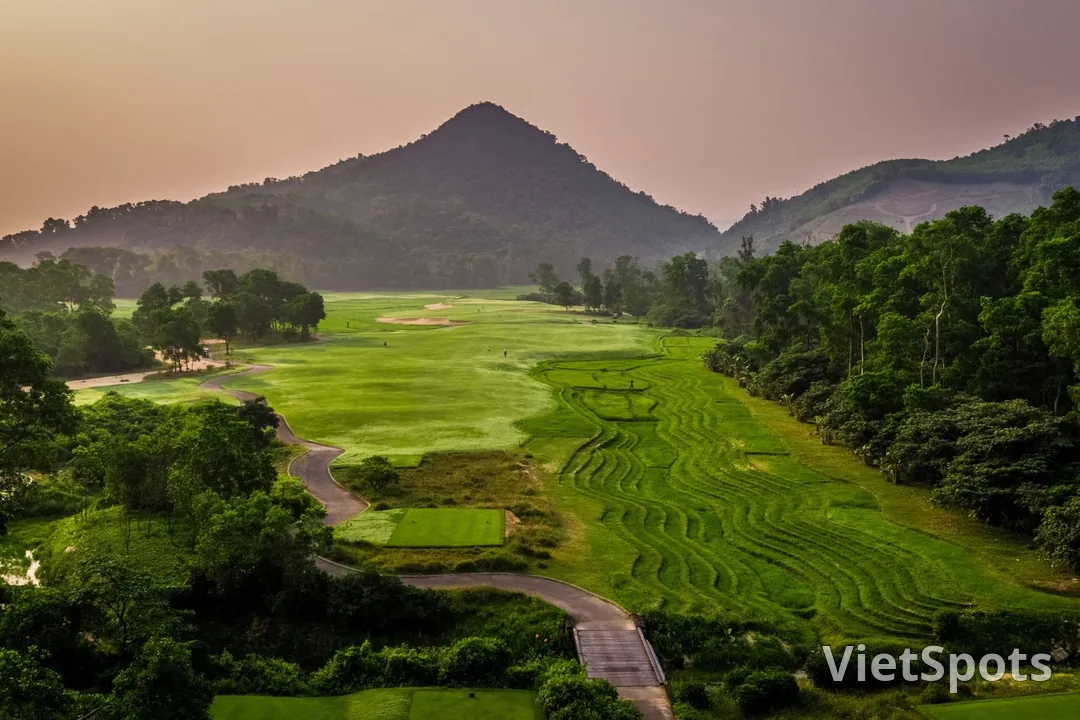 Fairway uốn qua thung lũng dưới một ngọn núi hình chóp, cỏ được cắt thành những đường cong như ruộng bậc thang