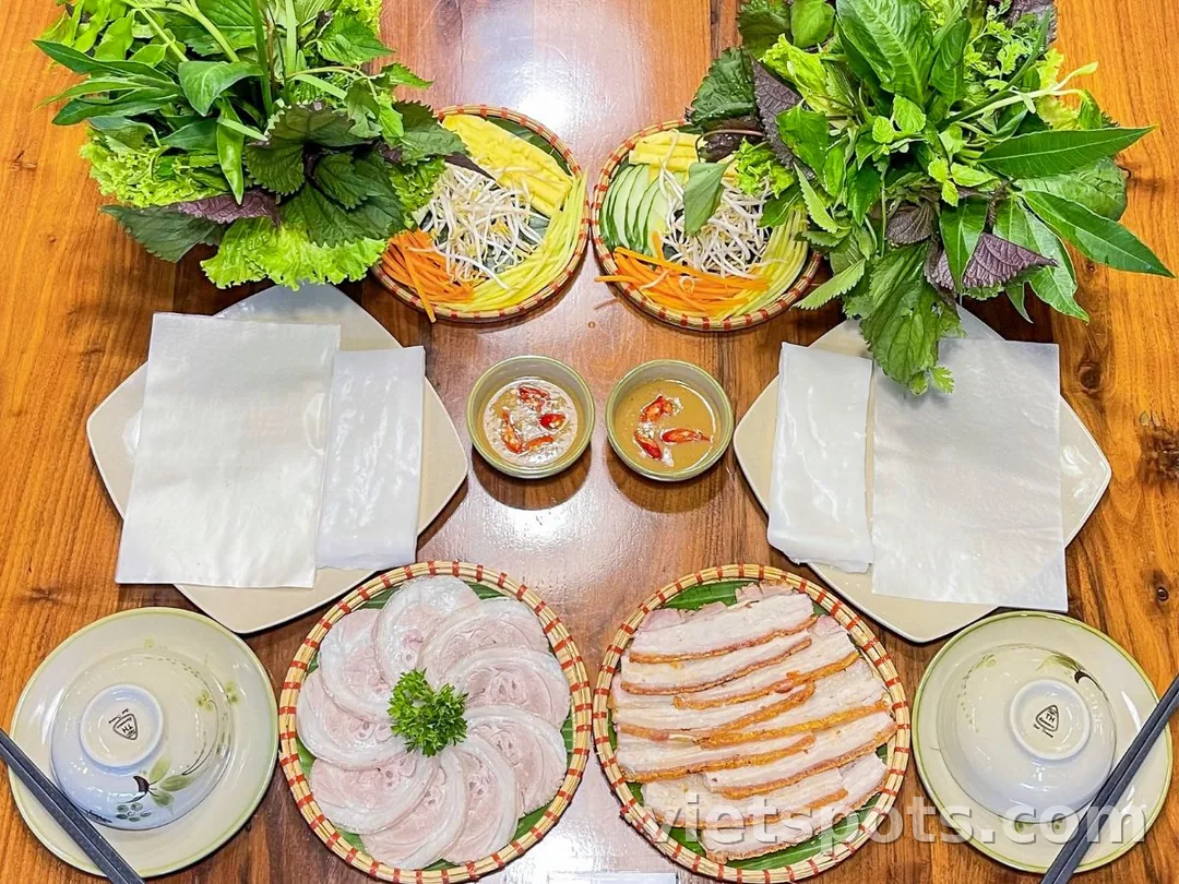 Bàn ăn bánh tráng cuốn thịt heo bày đối xứng — hai đĩa thịt luộc và heo quay, hai rổ rau, hai đĩa bánh tráng, hai chén mắm nêm