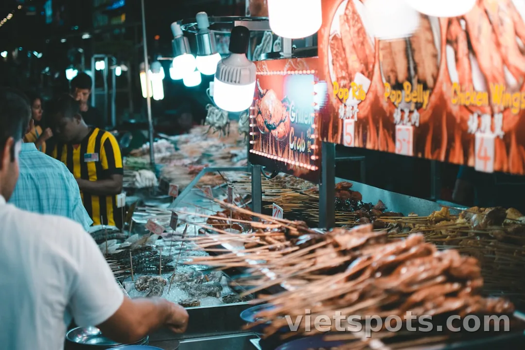 Night market châu Á — quầy BBQ skewers, đèn sáng, khách mua đêm