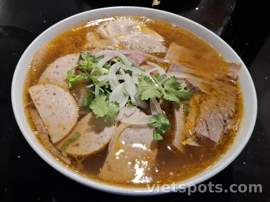 Tô bún bò Huế — nước dùng đỏ cam, thịt bò, chả cua, ngò hành trên