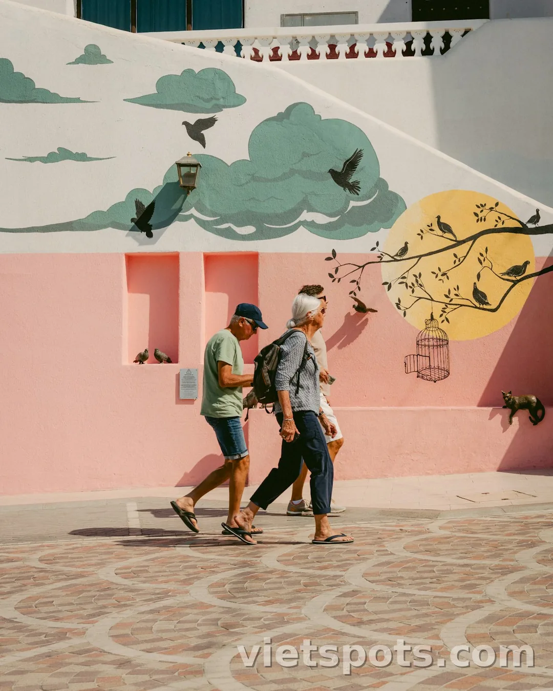 2 khách du lịch đi bộ ngang bức tường hồng có mural mây xanh, chim bay và lồng chim vàng