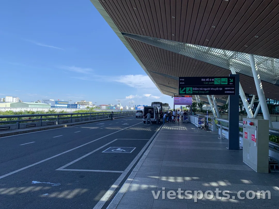 Cửa ra sân bay Đà Nẵng — mái gỗ hiện đại, biển hiệu Car park + PRM Drop-off tiếng Việt-Hàn-Anh-Trung, xe minivan đón khách