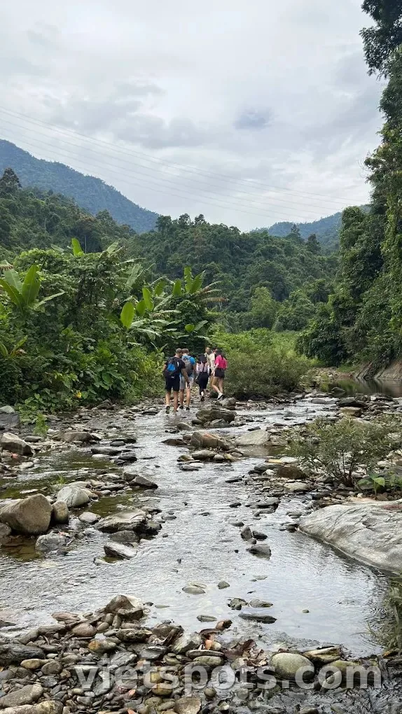 Nhóm 4-5 trekker balo đỏ tím xanh lội qua suối đá giữa rừng nhiệt đới, núi Bà Nà phía xa