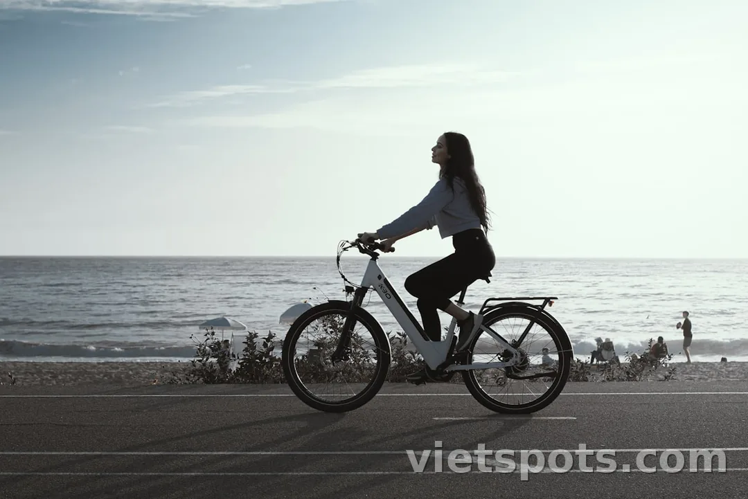 Em trẻ tóc dài mặc áo hoodie xám + quần đen casual, đạp xe city bike trắng trên đường ven biển, hoàng hôn nhẹ
