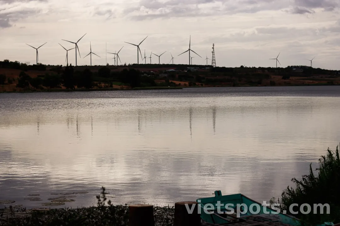 Hồ mặt phẳng lặng với hàng wind turbine phía xa, thuyền xanh nhỏ bên bờ