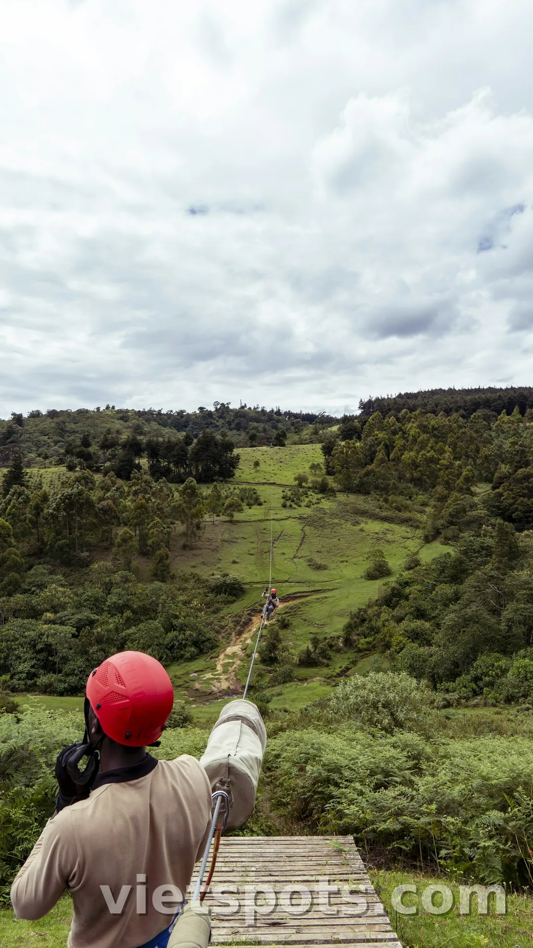Người đội mũ bảo hiểm đỏ chuẩn bị trượt zipline xuống thung lũng xanh, dây zip line dài phía trước
