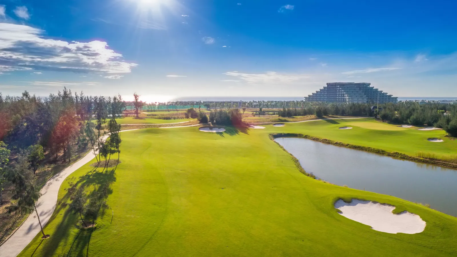 Một fairway rộng ở Vinpearl Golf Nam Hội An với hồ nước bên phải và rặng phi lao chắn gió ven biển