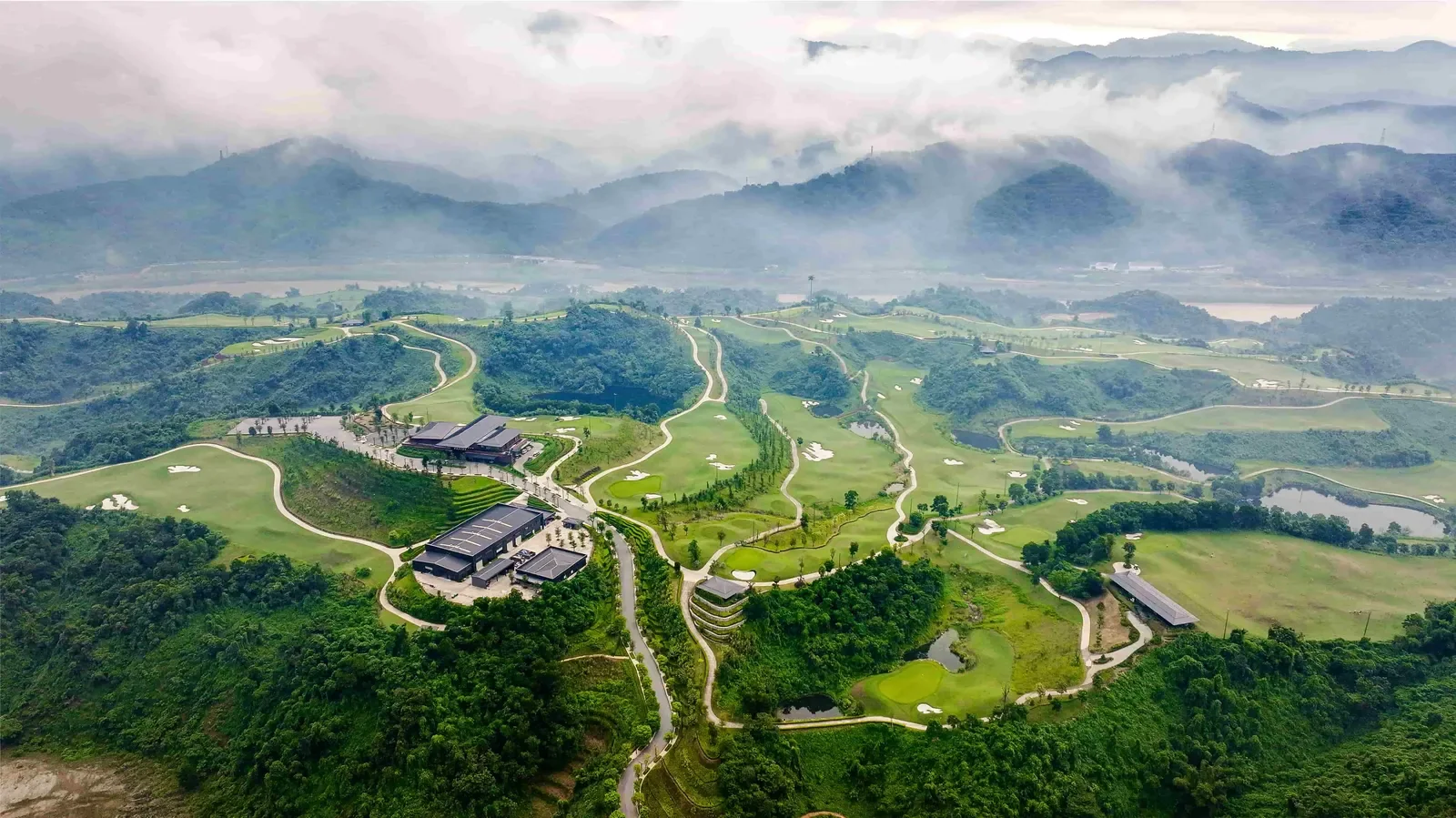 Sân Sapa Grand nhìn từ trên cao, các fairway uốn giữa đồi rừng với mây thấp phủ kín dãy núi phía sau
