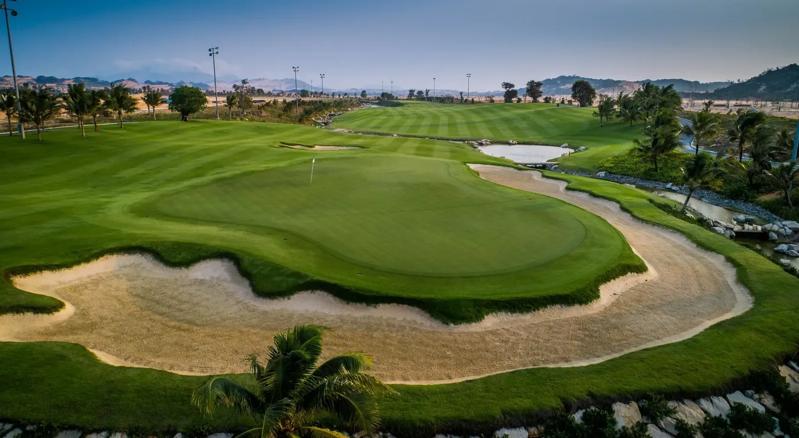 Green ở KN Golf Links Cam Ranh với bẫy cát lớn bao quanh, phía sau là đồi trọc vùng Ninh Thuận lúc chiều muộn